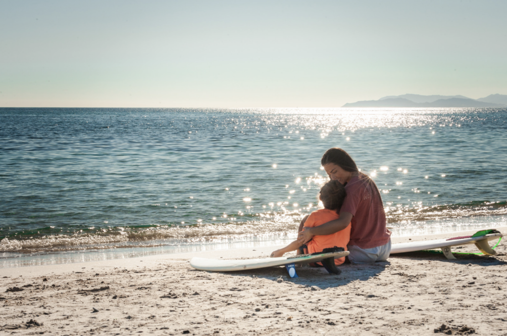 This is an image of a mother and child sitting together on the beach with some surfboards nearby.